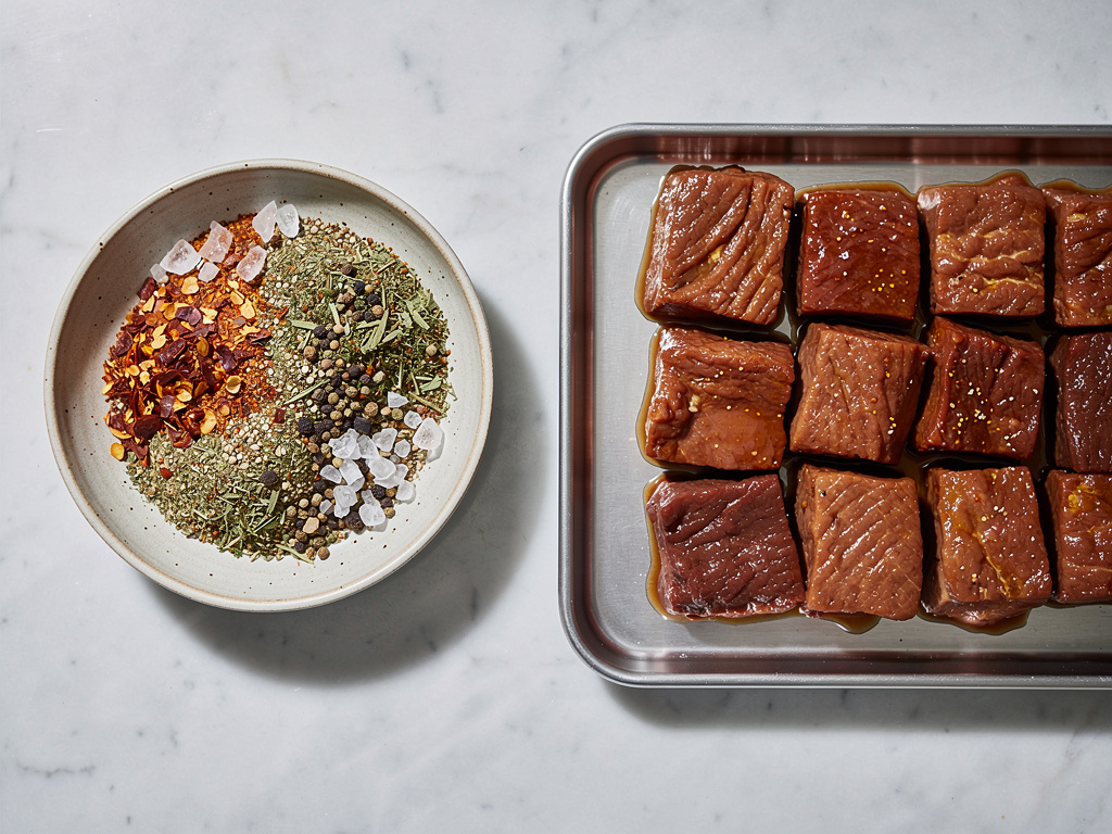 A bowl of coarse seasoning and herbs beside marinated meat pieces in a shallow tray on marble.